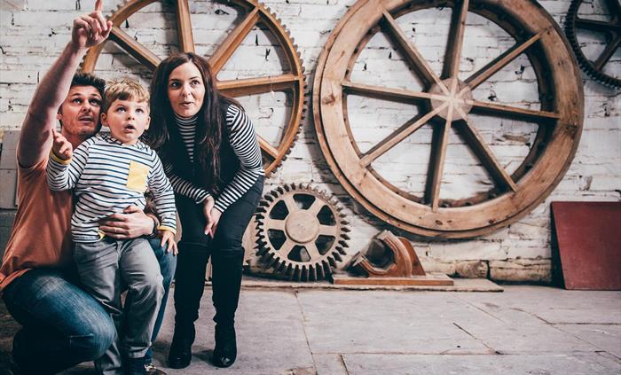 two parents and young son explore the pattern loft. There are wooden models, known as patterns, of wheels and cogs. These patterns were used to cast w